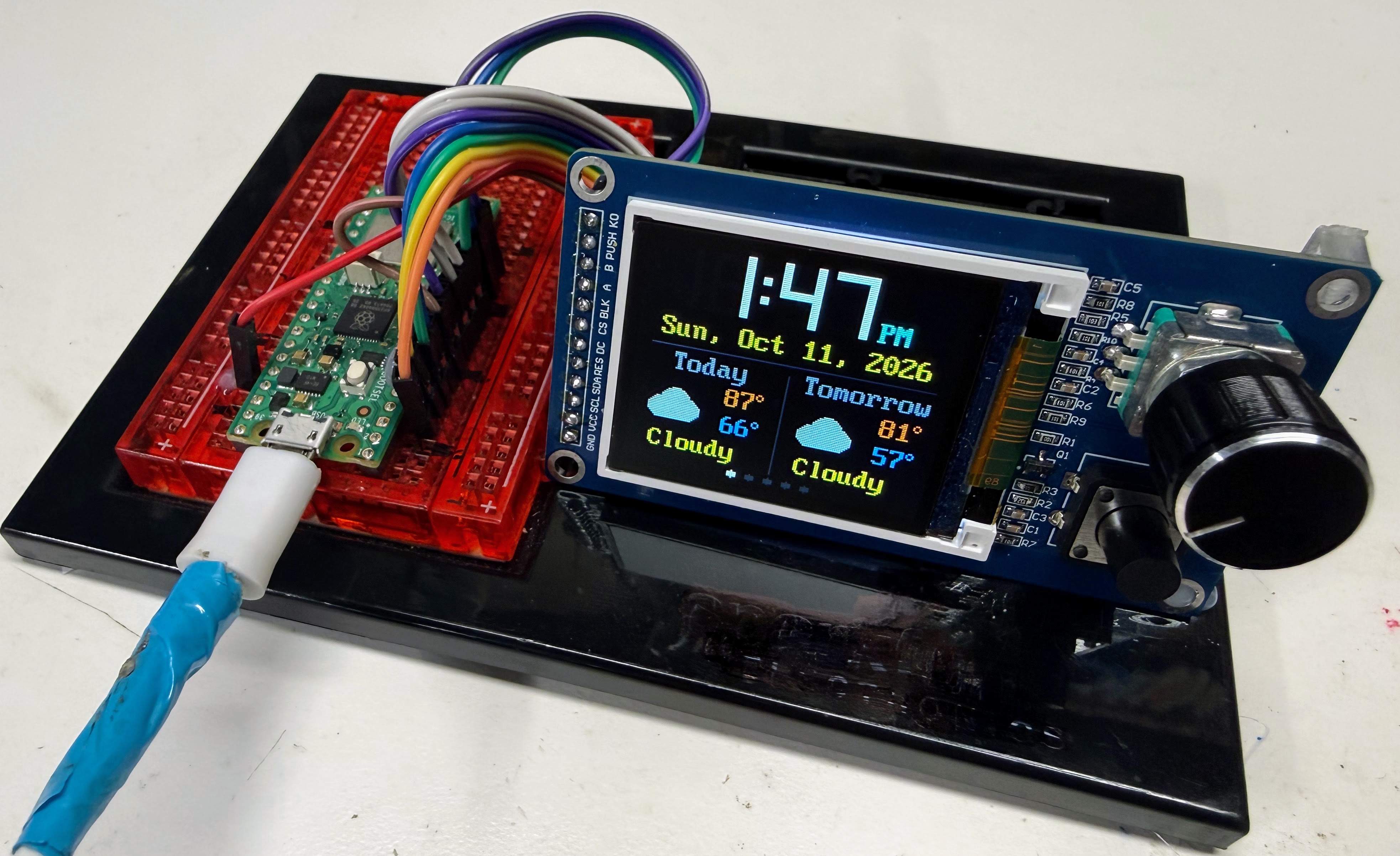 The Knob Clock Kit on a breadboard: a Raspberry Pi Pico 2 W joined by a rainbow ribbon cable to a small rectangular color screen. The screen shows the weather clock: 1:47 PM, Sunday, October 11, 2026, cloudy today with a high of 87 and a low of 66, cloudy tomorrow with 81 and 57, and a row of five dots along the bottom. A round black knob and a square push button sit to the right of the screen.