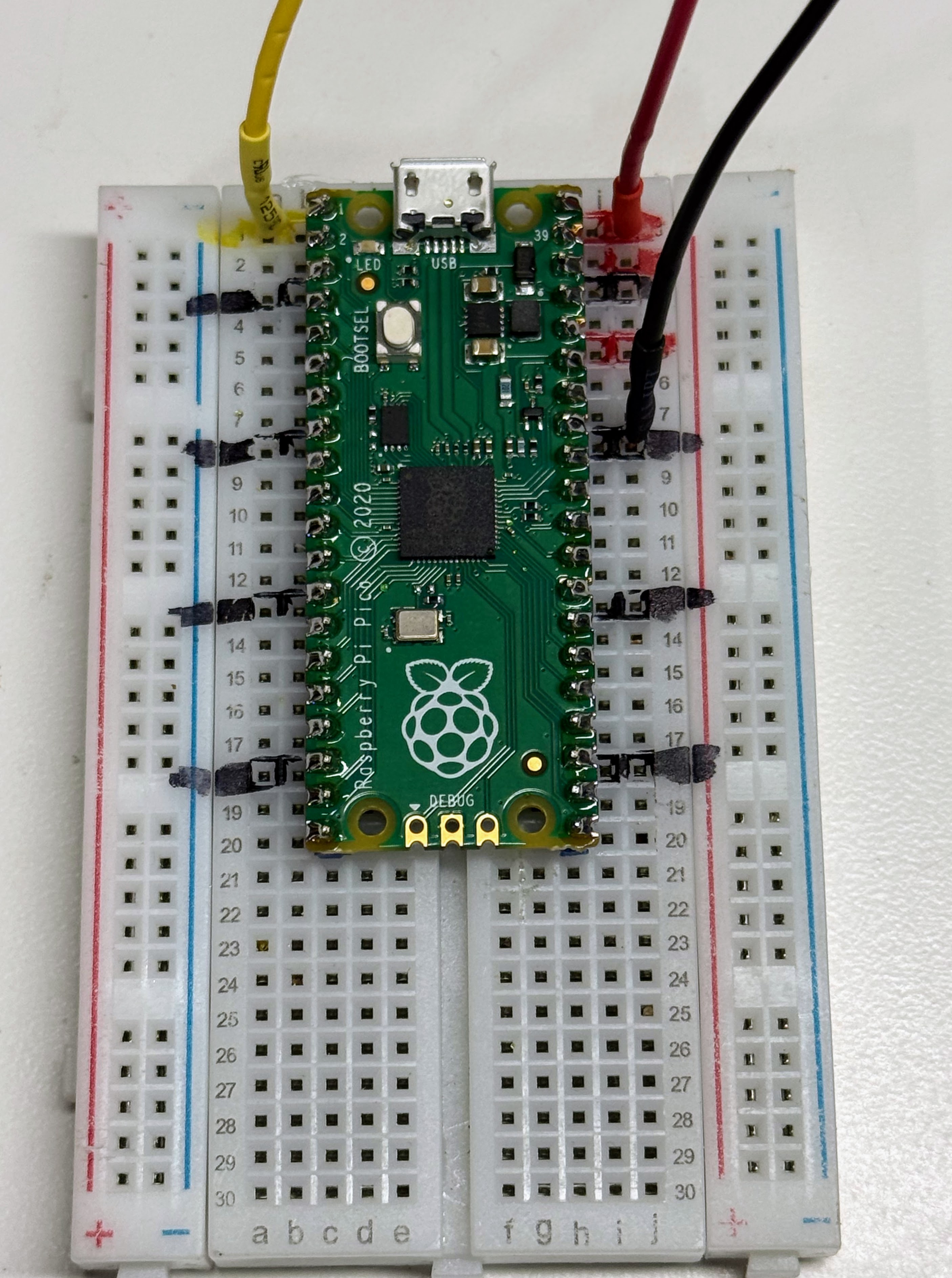 A Raspberry Pi Pico seated on a breadboard with the USB connector at the top, showing the yellow data wire at GP0, the red power wire at VBUS, and the black ground wire at a GND pin